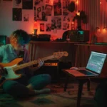 A young musician with colorful hair plays a bass guitar in a vibrant, messy bedroom studio with a laptop and lava lamp.